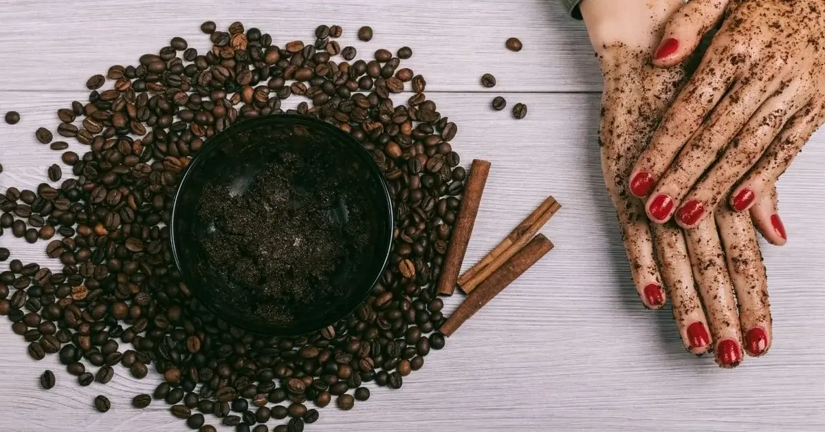 Scrub z kawy w miseczce, rozsypane ziarna kawy i dłonie nasmarowane peelingiem kawowym