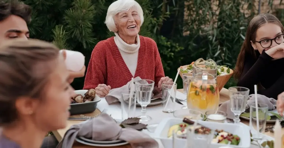 Rodzina ze starszą kobietą w czerwony kardiganie w radosnym nastroju siedzi dookoła stołu z białym serwisem obiadowym