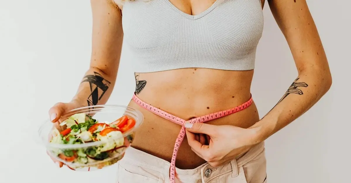 Sylwetka kobiety w jasnym topie, w ręku trzyma miskę z warzywami, drugą ręką mierzy centymetrem obwód w talii