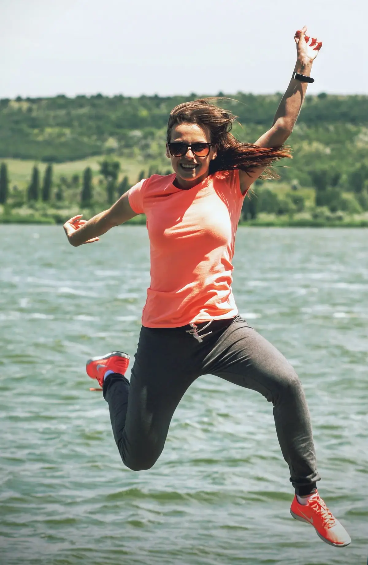 Radośnie skacząca w górę kobieta ubrana w sportową pomarańczową koszulkę, dresowe spodnie i sportowe buty na tle jeziora