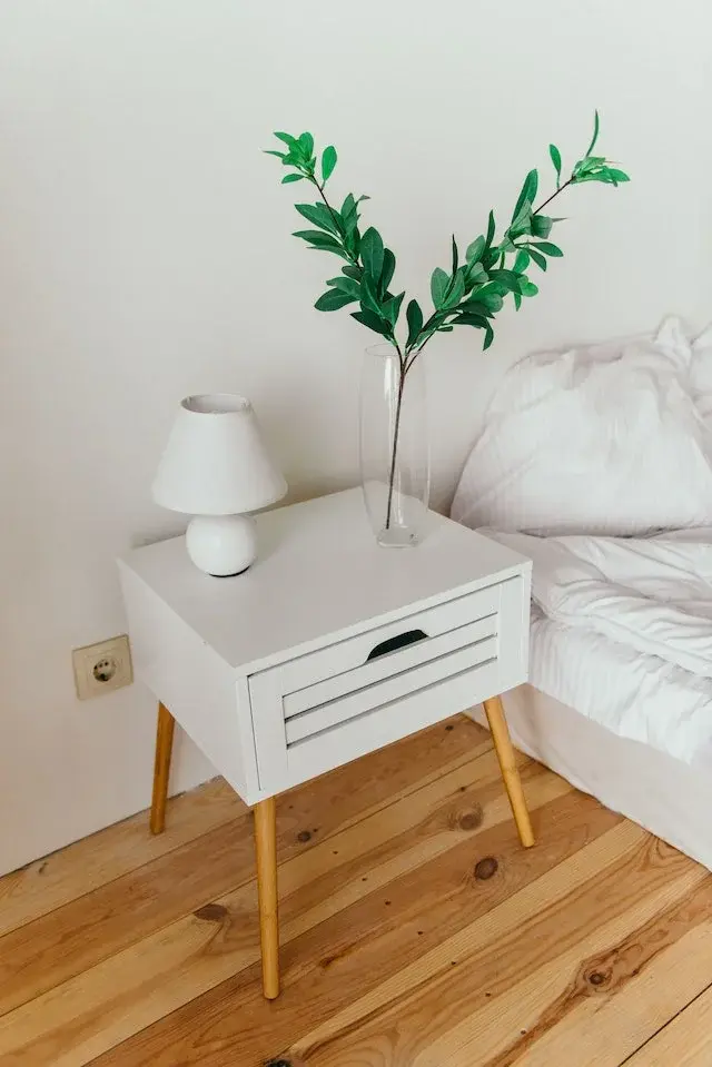 Biały stolik nocny z drewnianymi nóżkami na drewnianej podłodze obok białego łóżka