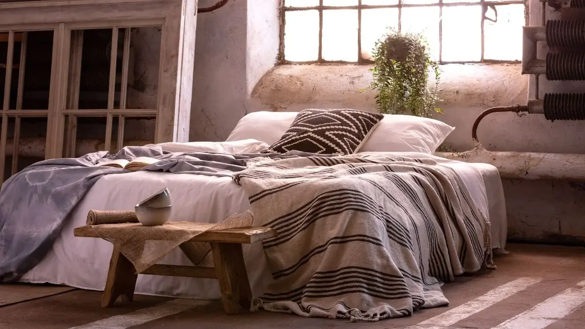Sypialnia w stylu wabi-sabi z łóżkiem przykrytym naturalnymi narzutami, drewnianą ławką w nogach, na której stoją czarki