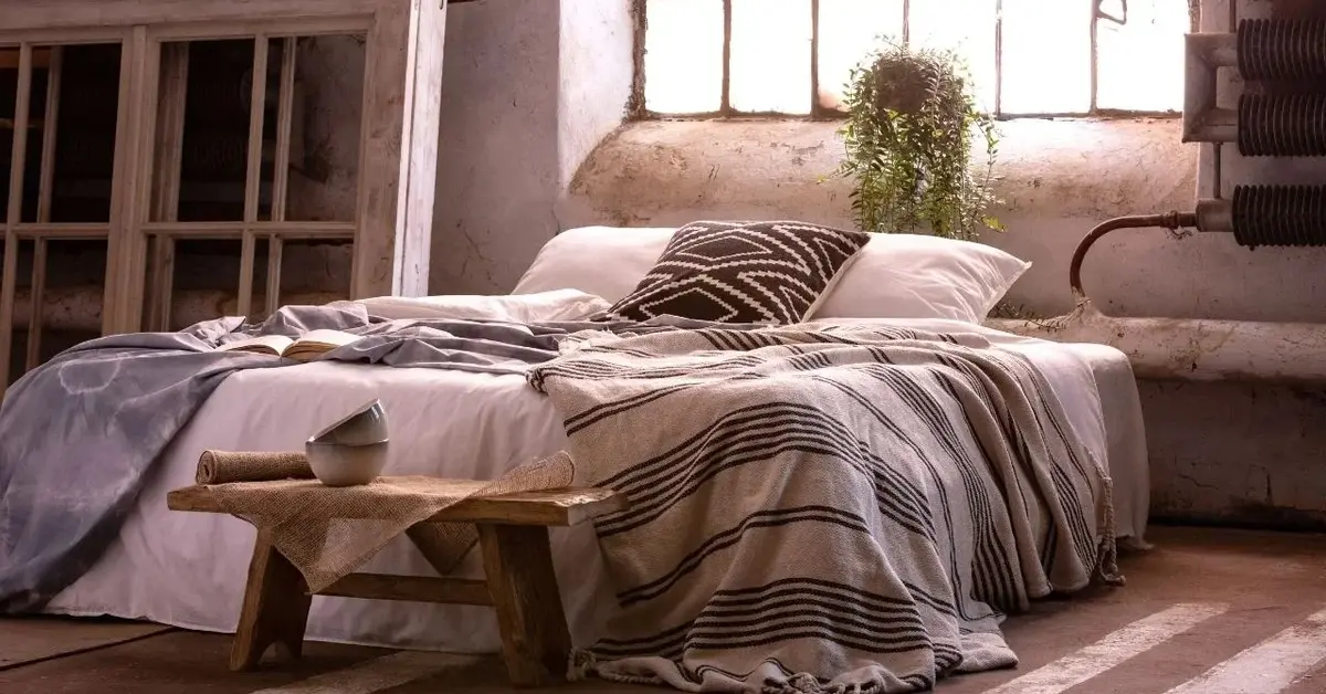 Sypialnia w stylu wabi-sabi z łóżkiem przykrytym naturalnymi narzutami, drewnianą ławką w nogach, na której stoją czarki