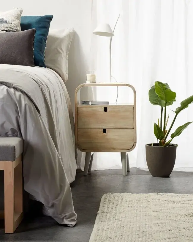 Drewniana szafeczka nocna z białą lampką stoi przy zaścielonym łóżku, bok niej kwiat w doniczce