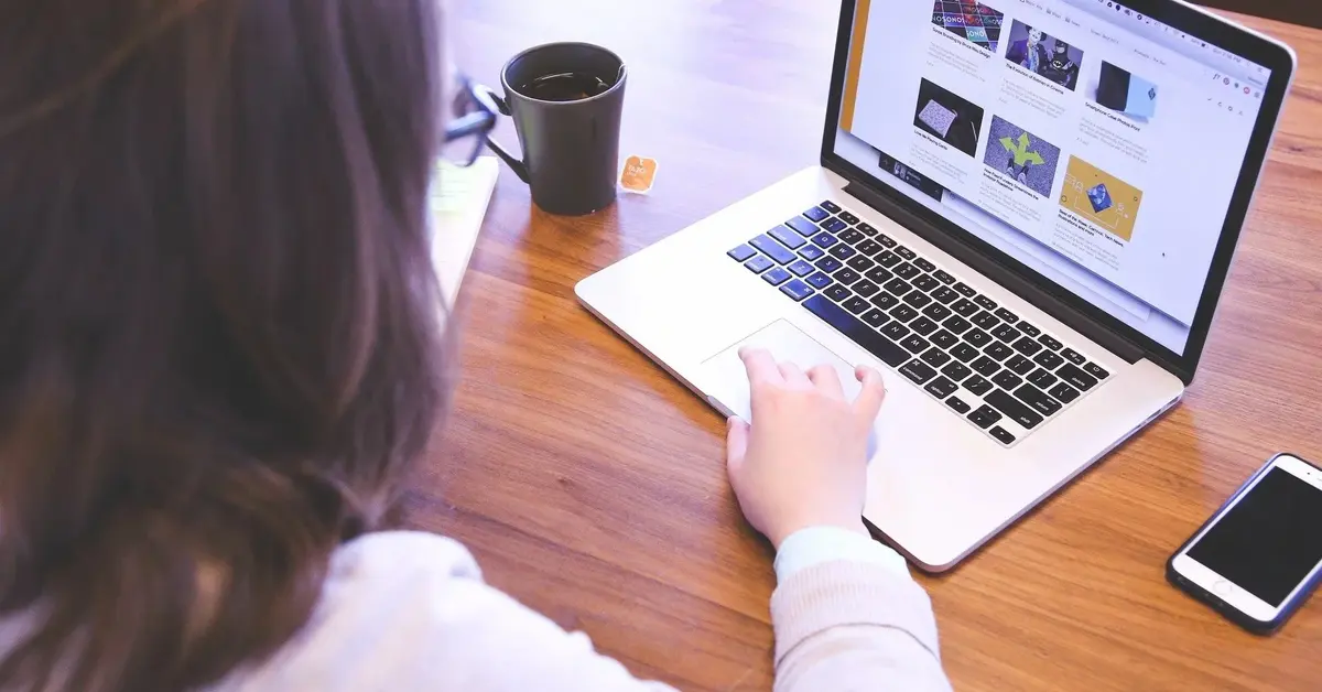 Przy brązowym biurku kobieta brązowe włosy w białej koszuli widziana od tyłu szuka w laptopie pracy przez internet
