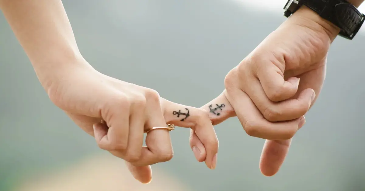 Kobieta i mężczyzna mają splecione palce wskazujące, na których mają tatuaże kotwicy