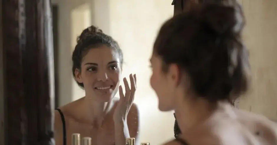 Kobieta w domowym koku i koszulce na ramiączkach przed lustrem w trakcie tonizowania twarzy jako etapy pielęgnacji cery
