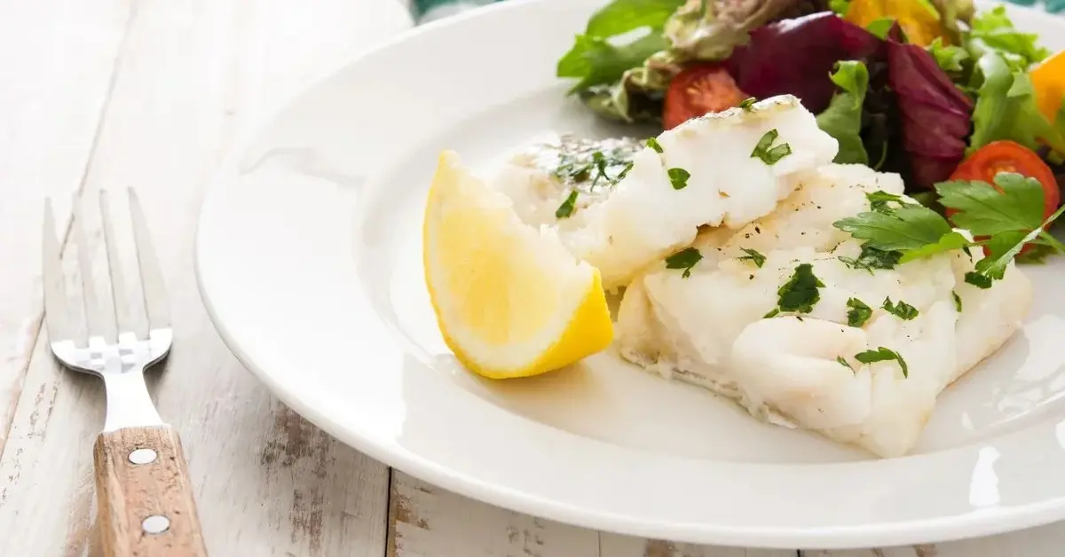 Na stole na białym talerzy danie z białej ryby morskiej trewal z kawałkiem cytryny posypane ziołami, obok widelec