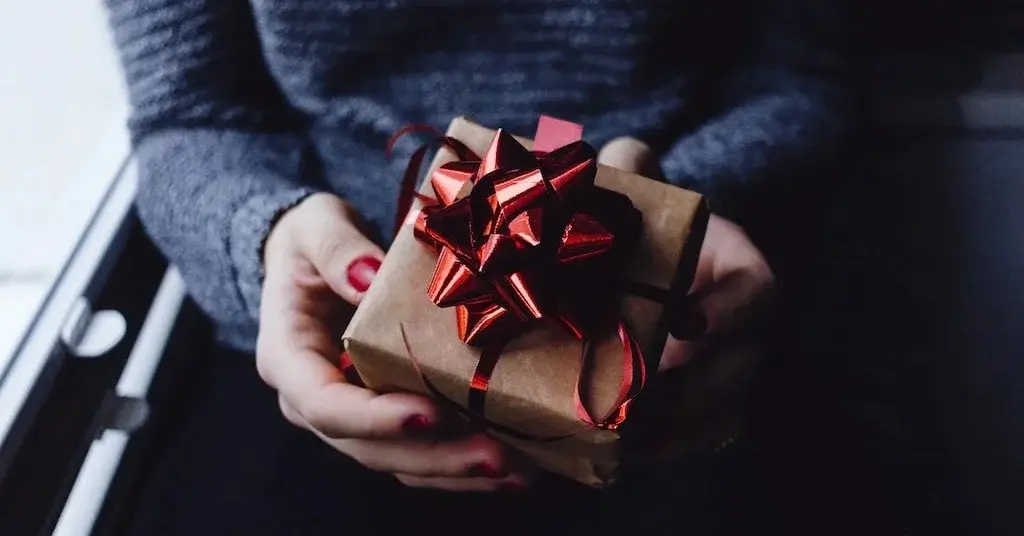 Kobiece dłonie o czerwonym manicure trzymające drobny uniwersalny top prezent na Mikołajki z czerwoną koakrdą