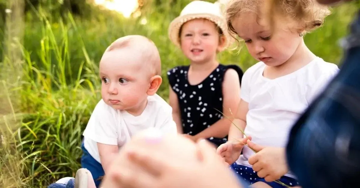 W czasie urodzin dziecka w plenerze grupa dzieci w różnym wieku siedzi na trawie z radosnymi minami