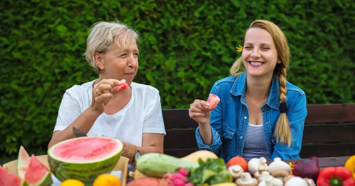 owoce i warzywa na stole z tyłu dwie kobiety śmieją się i jedzą za nimi tło z roślin