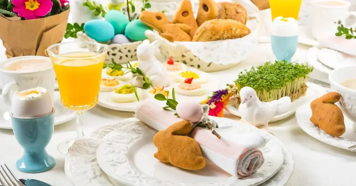 stół zastawiony talerze serwetki ciasteczka w kształcie królików jajka turkusowe kwiaty różowe trawka sok pomarańczowy 