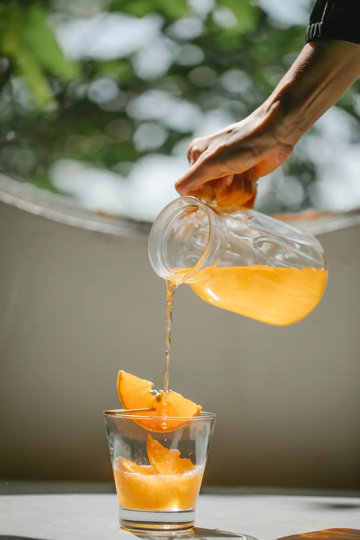 Dłoń nalewająca ze szklanego dzbanka do szklanki z pomarańczą sok pomarańczowy z witaminą C na tle okna