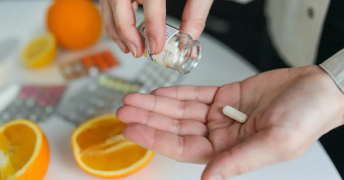 Ludzkie dłonie wytrząsające ze słoiczka tabletkę w ramach suplementacji witaminy C na tle stołu z pomarańczami
