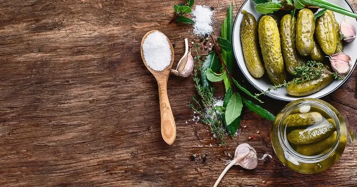 Na drewnianym blacie widok z góry na talerz pełen ogórków, obok słoik z ogórkami kiszonymi w zalewie, zioła, przyprawy