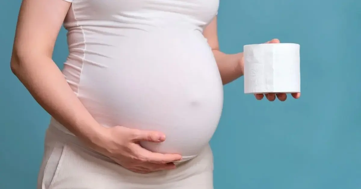 Brzuch ciężarnej kobiety w jasnej sukience, ona trzyma w dłoni rolkę papieru toaletowego z powodu zaparć w ciąży