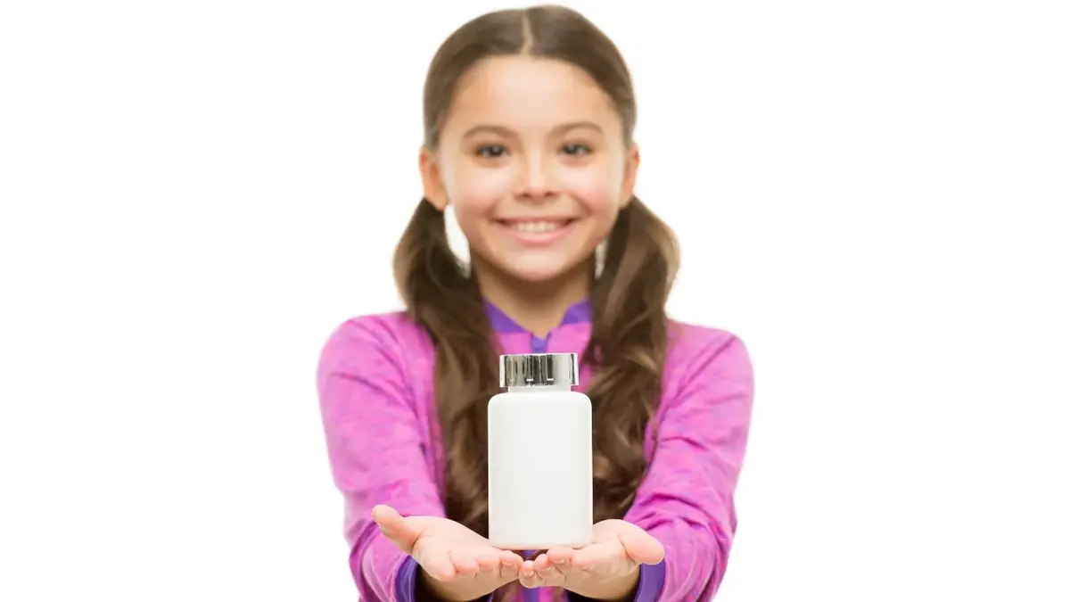 Dziewczynka w fioletowej bluzeczce trzyma przed sobą białą buteleczke z suplementami