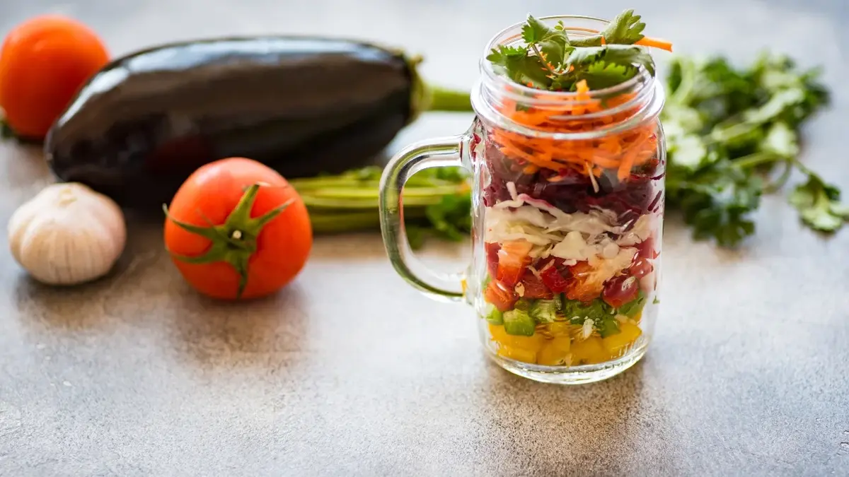 Sałatka w słoiku ze świeżymi warzywami. Obok warzywa: pomidor, bakłażan, czosnek i pietruszka