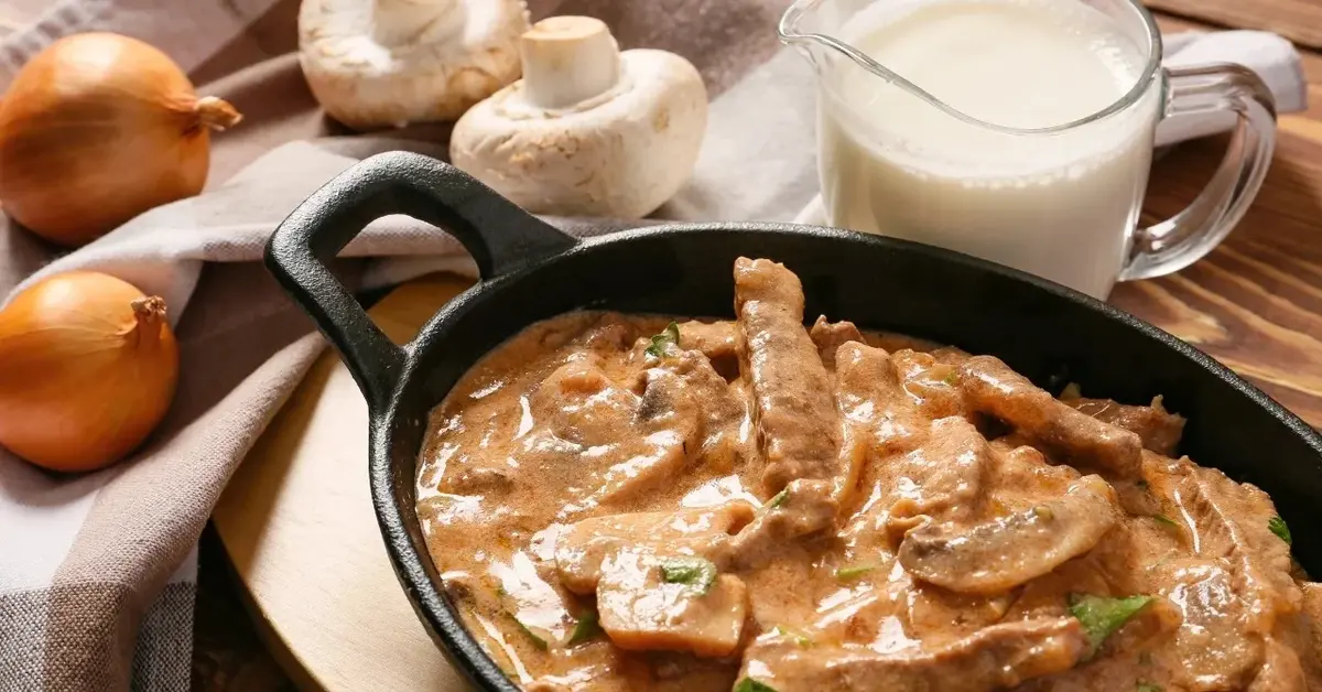 Żeliwne naczynie wypełnione strogonowem z kawałkami mięsa. Obok cebula, pieczarki i dzbanuszek ze śmietaną