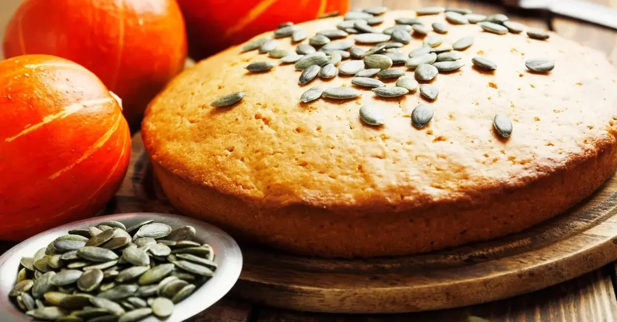 Okrągłe ciasto dyniowe posypane ziarenkami dyni. Obok leżą trzy małe ozdobne dynie i mały talerzyk z nasionami dyni