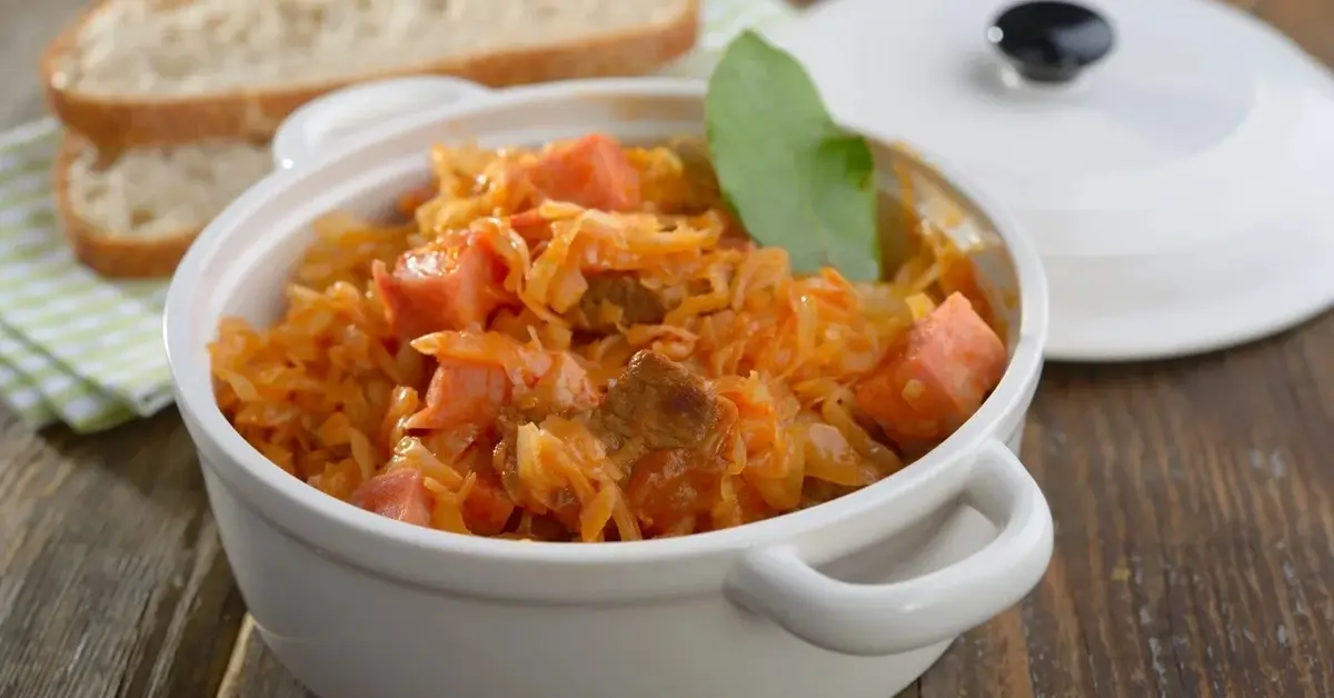 Bigos w białym małym garnku, obok biała pokrywka w tle dwie kromki chleba