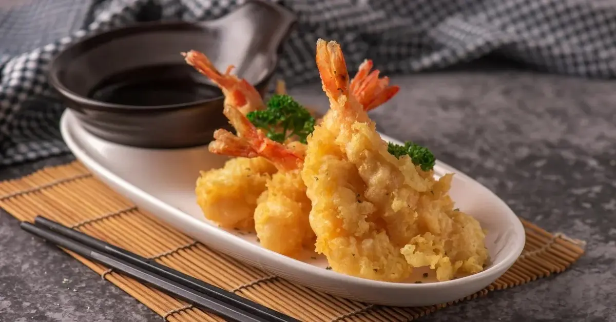 Krewetki w panierce na białym półmisku stojącym na macie bambusowej. Obok pałeczki