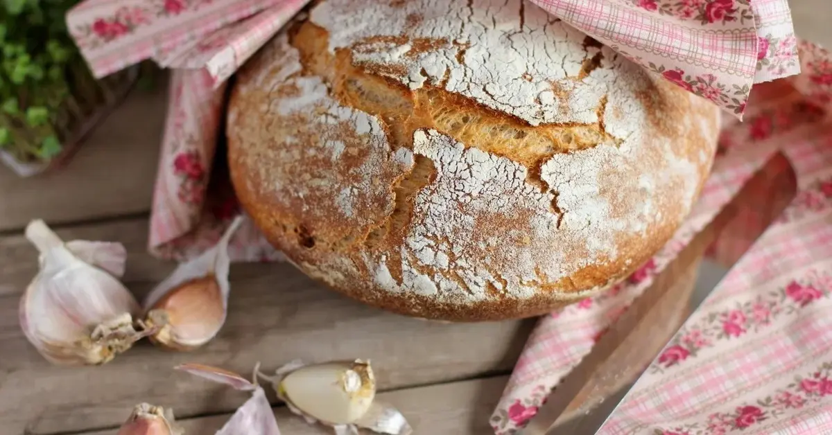 Chleb domowy w połowie owinięty serwetką. na blacie rozrzucone ząbki czosnku i nóż