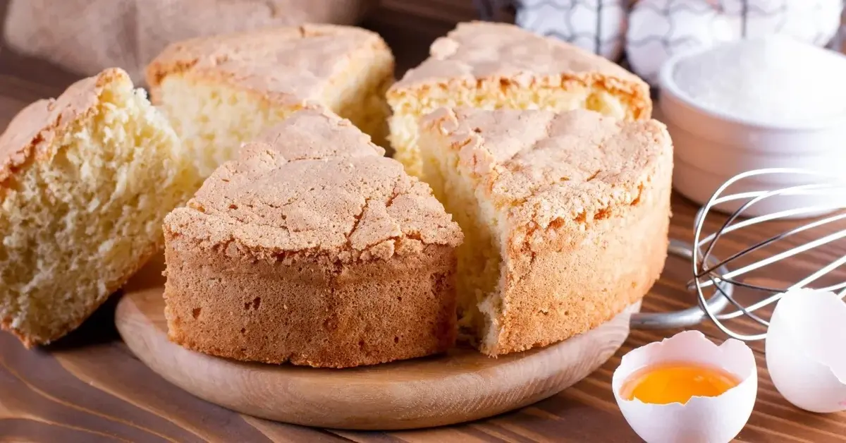 Biszkopt okrągły pokrojony na kawałki ułożony na drewnianej desce. Obok przybory kuchenne i jajko