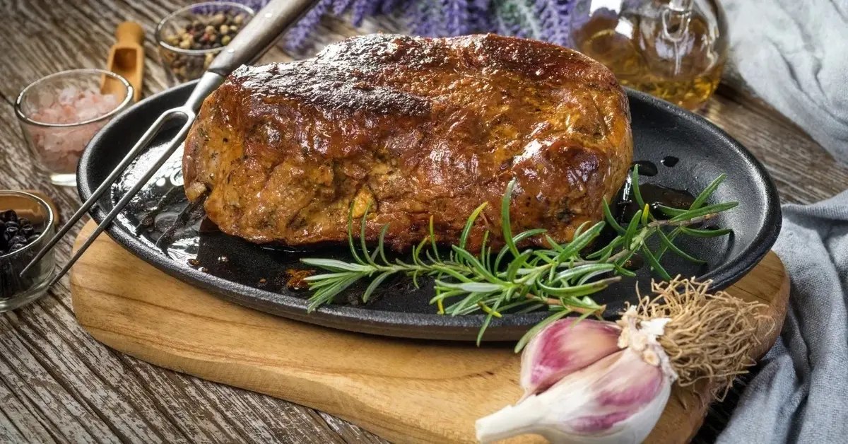 KArkóka pieczona w kawałku na talerzu i drewnianej desce. Obok rozmaryn i główka czosnku