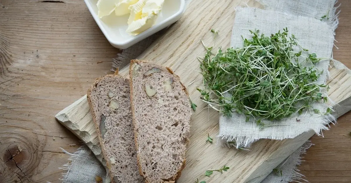 Dwie kromki chlebka ziołowego na drwnianej desce. Obok maselniczka z masłem i świeże zioła