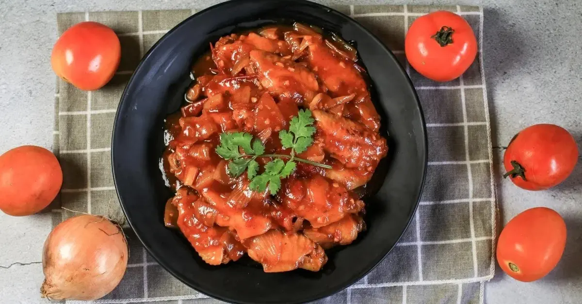 Gulasz z indyka w sosie pomidorowym na czarnym talerzu. Obok pomidory i cebula