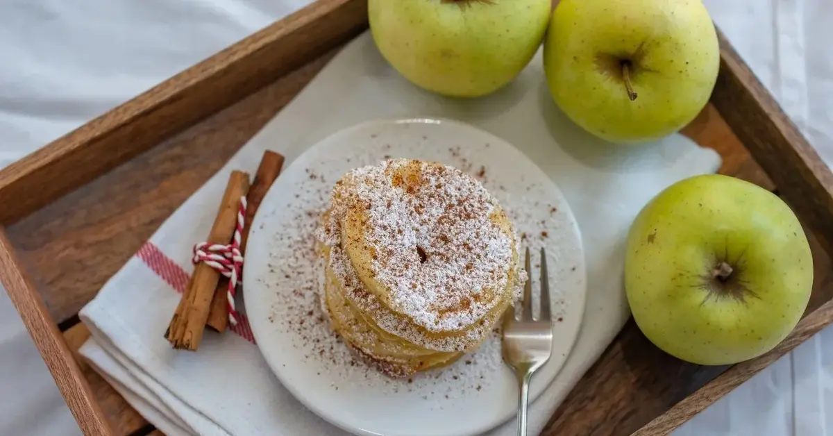 Na drewnianej tacy talerzyk z racuchami z jabłkami, obok trzy zielone jabłka