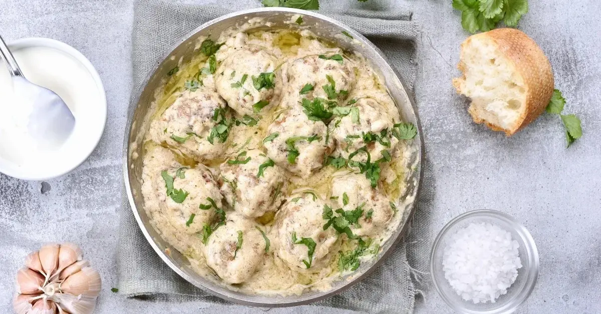 Pulpety w sosie koperkowym w garnku, obok miseczka ze śmietaną, kawałek bułki i czosnek