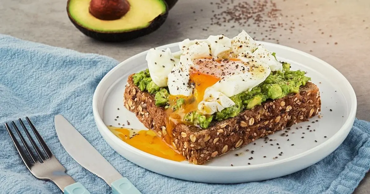 Tost z jajkiem w koszulce na białym talerzyku, obok awokado