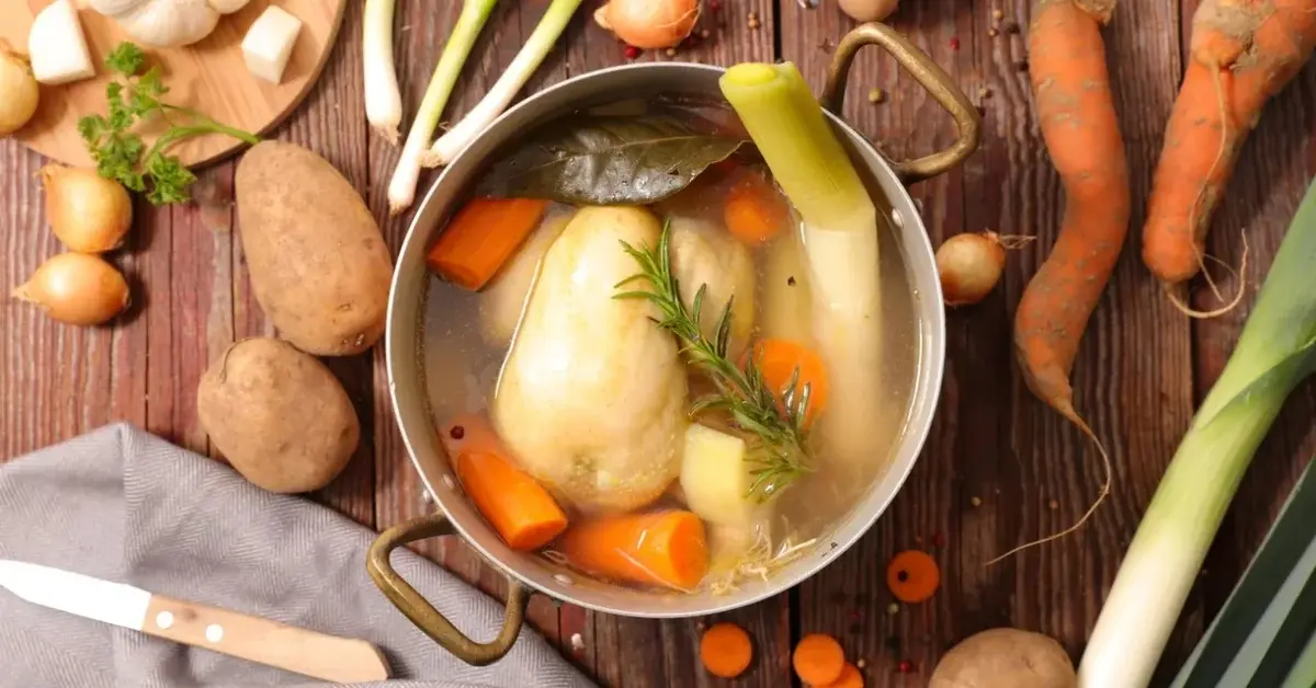 W dużym garnku rosół z warzywami i mięsem kurczaka, obok na blacie warzywa: marchewki, ziemniaki, cebulki, por