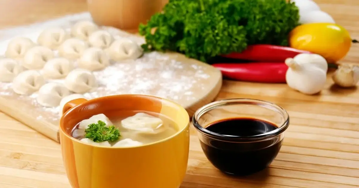 Zupa won ton w bambusowej miseczce, obok miseczka z sosem sojowym i pierożki won ton na drewnianej desce