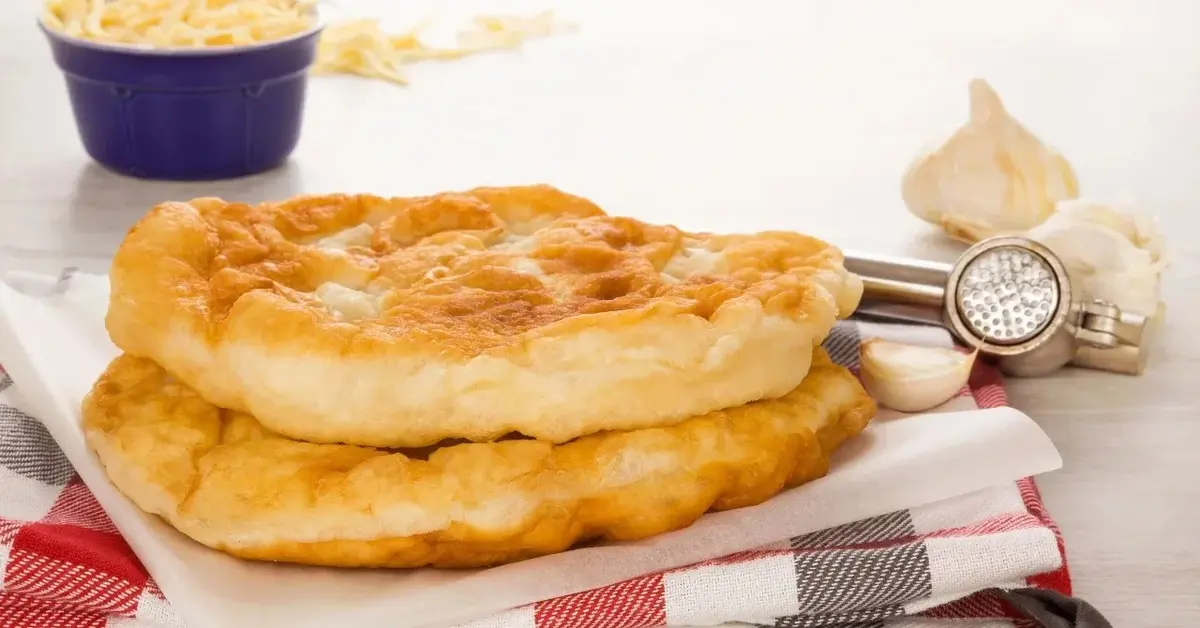 Langosz ze śmietaną, obok główki czosnku, wyciskarka do czosnku i tarty ser w miseczce