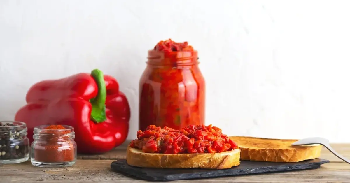 Pasta ajwar na kromce chleba, obok słoik z pastą i papryka czerwona