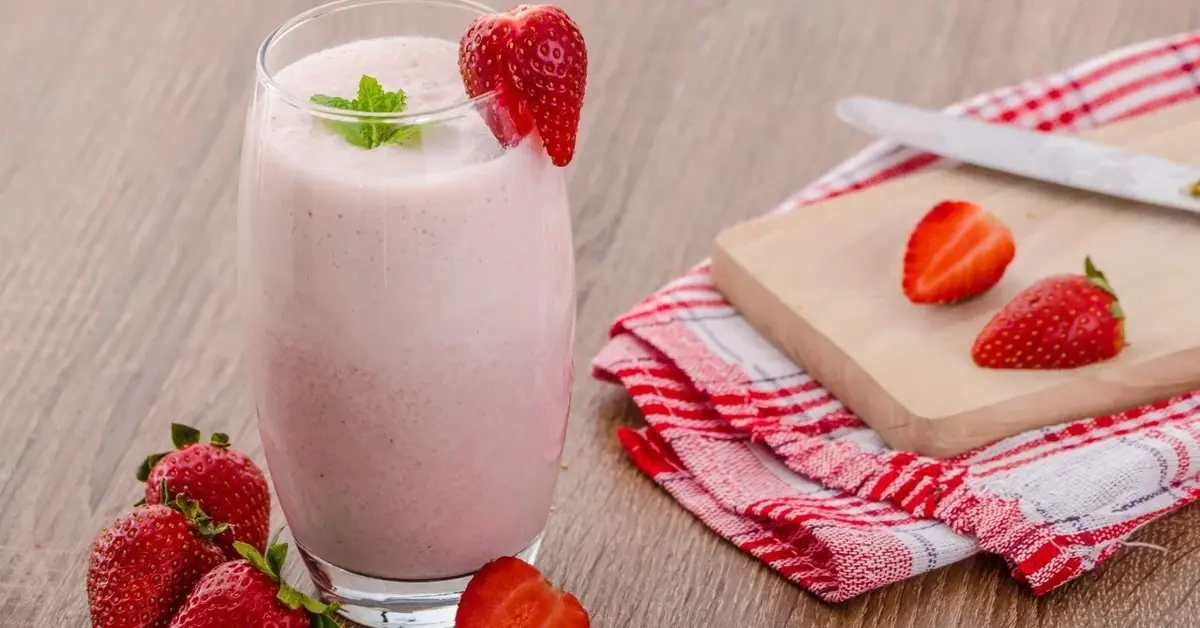 Smoothie truskawkowe w szklance, obok drewniana deska do krojenia i truskawki