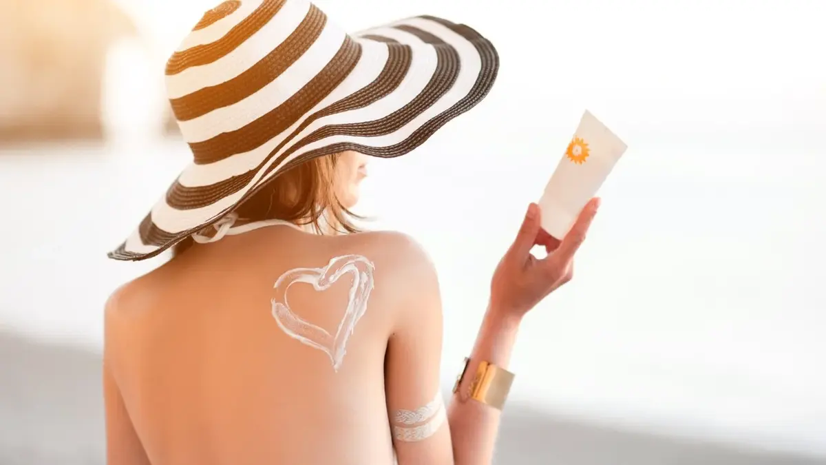 Kobieta w kapeluszu z nałożonym kremem przeciwsłonecznym, którego opakowanie trzyma w ręce