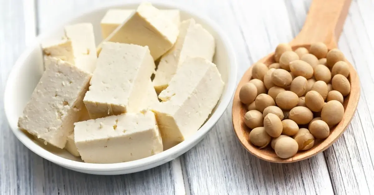 Tofu pokrojone w plasterki na półmisku. Obok drewniana łyżka z ziarnami soi