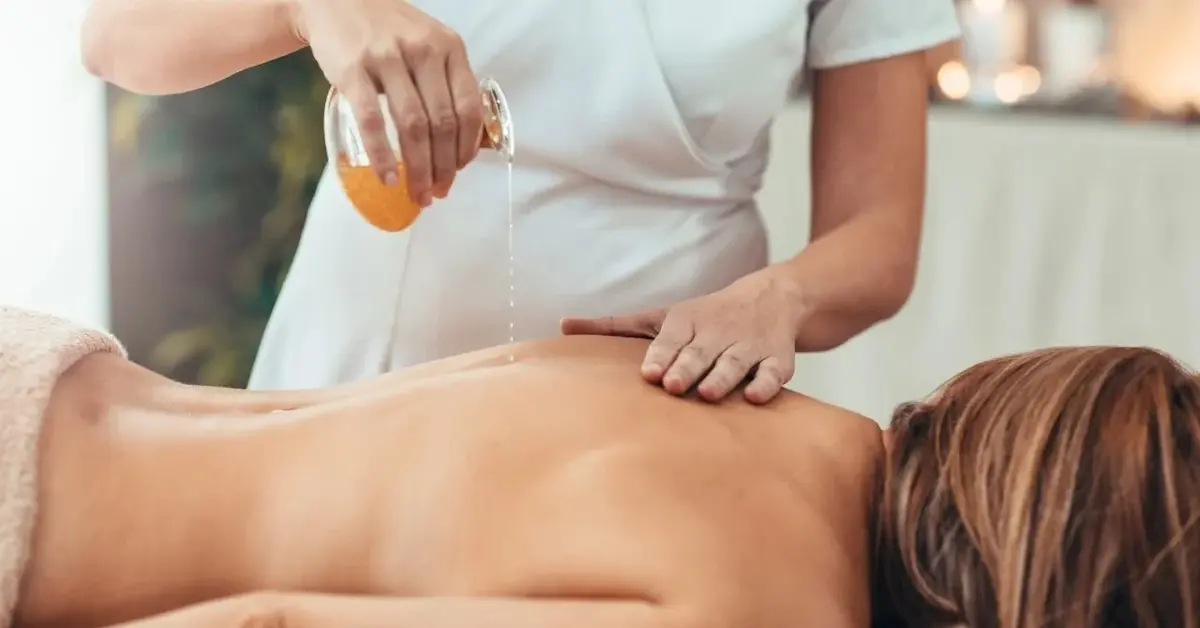 Kobieta na stole do masażu, obok druga osoba przygotowująca ją do masażu wylewa na plecy olejek