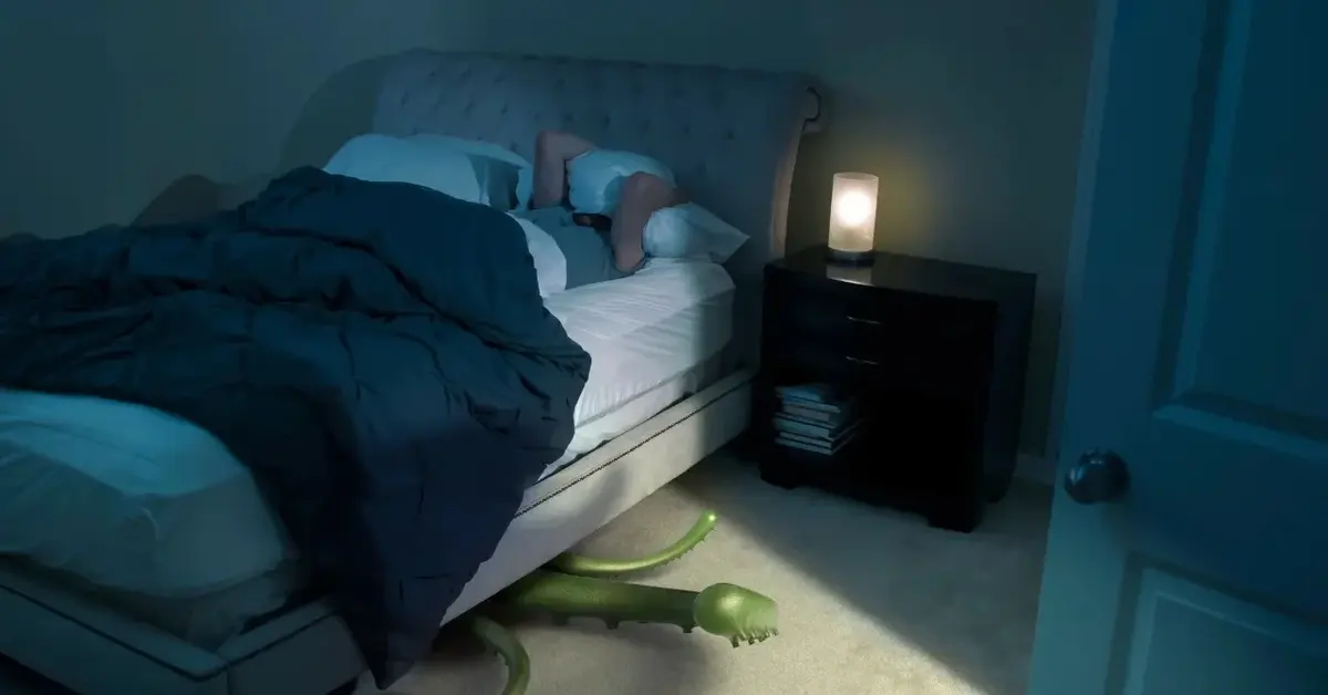 Mężczyzna leżący w łóżku z głową przykrytą poduszką. Spod łóżka wysuwa się zielony potwór