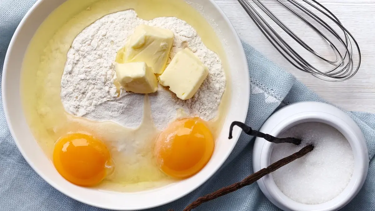 W białej misce nasypana jest mąka, kawałki masła i dwa rozbite jajka. Obok biała cukierniczka z dwiema laskami wanilii 