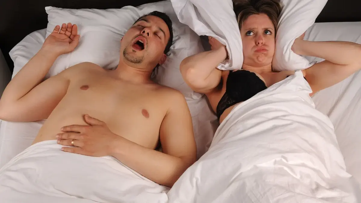 Kobieta leżąca w łóżku zasłaniająca uszy poduszką, obok śpiący mężczyzna