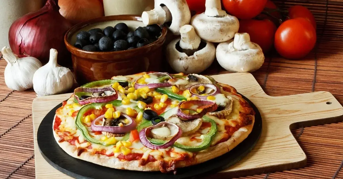Pizza z warzywnymi dodatkami na okrągłym blacie do pieczenia pizzy stojącym na drewnianej desce. W tle warzywa 