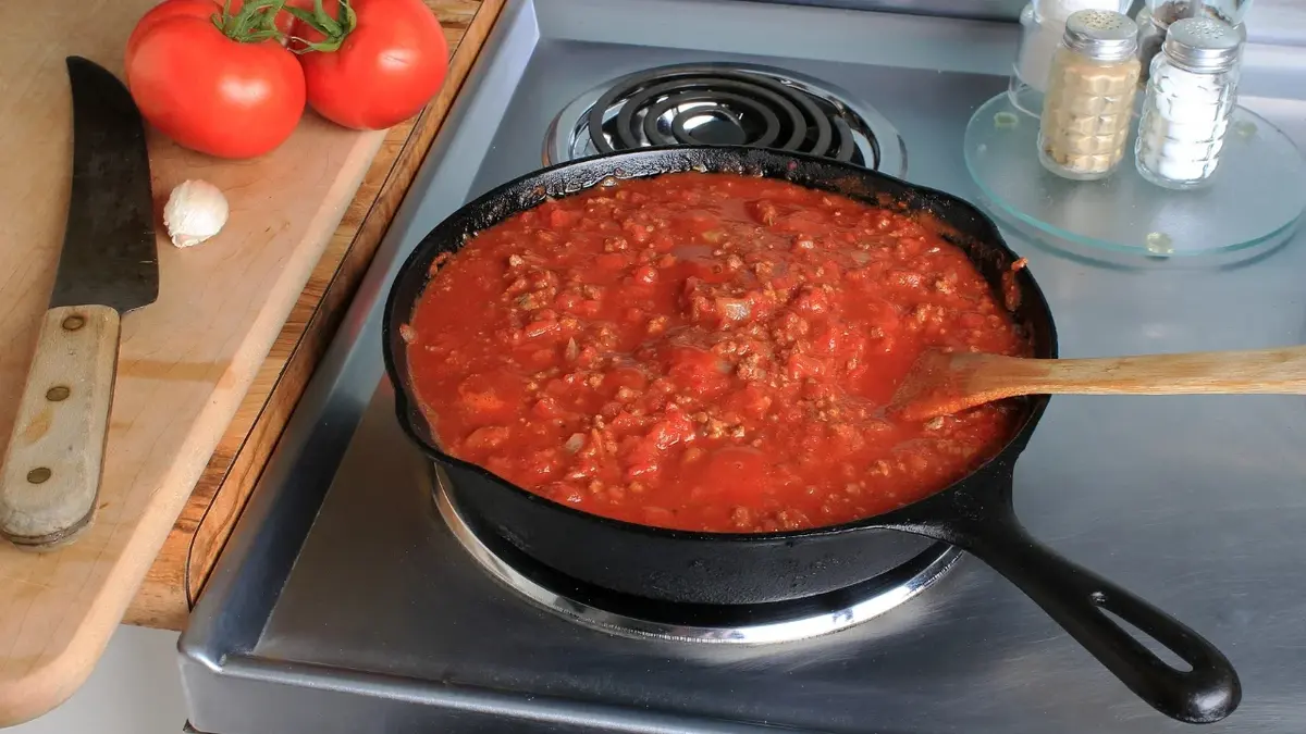 Sos do spaghetti w czarnej patelni na kuchence. obok nóż, pomidory i pieprz i sól