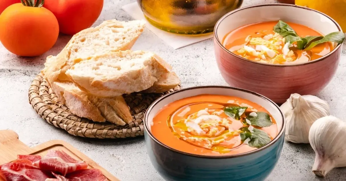 Zupa pomidorowa po hiszpańsku w dwóch miseczkach. Obok kromki bagietki na podstawce. W tle pomidory i główki czosnku