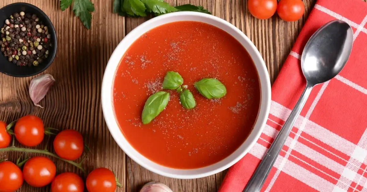 Zupa pomidorowa w białej miseczce na drewnianym blacie. Obok serwetka w biało-czerwoną kratę, pomidorki, łyżka, bazylia