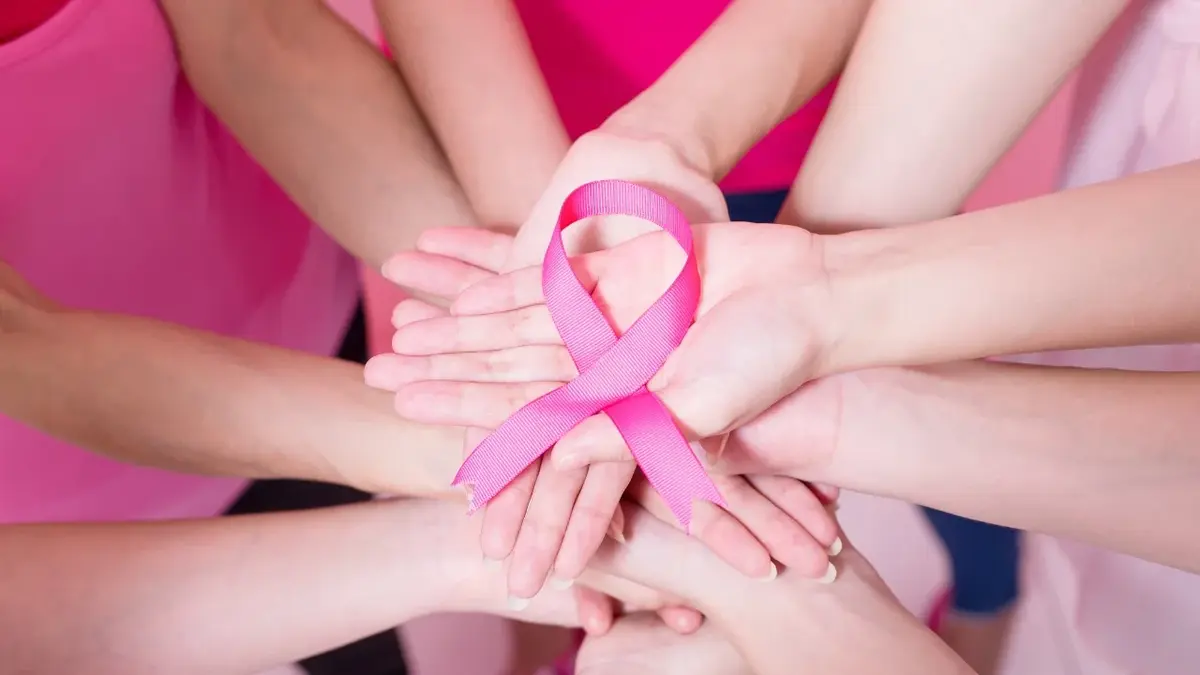 Ręce kobiet złączone na środku okręgu, na nich różowa wstążka w kształcie symbolu walki z rakiem piersi 